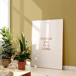 “Focus on the Lord” minimalist Christian wall art in lightwood frame beside plants, instant download.